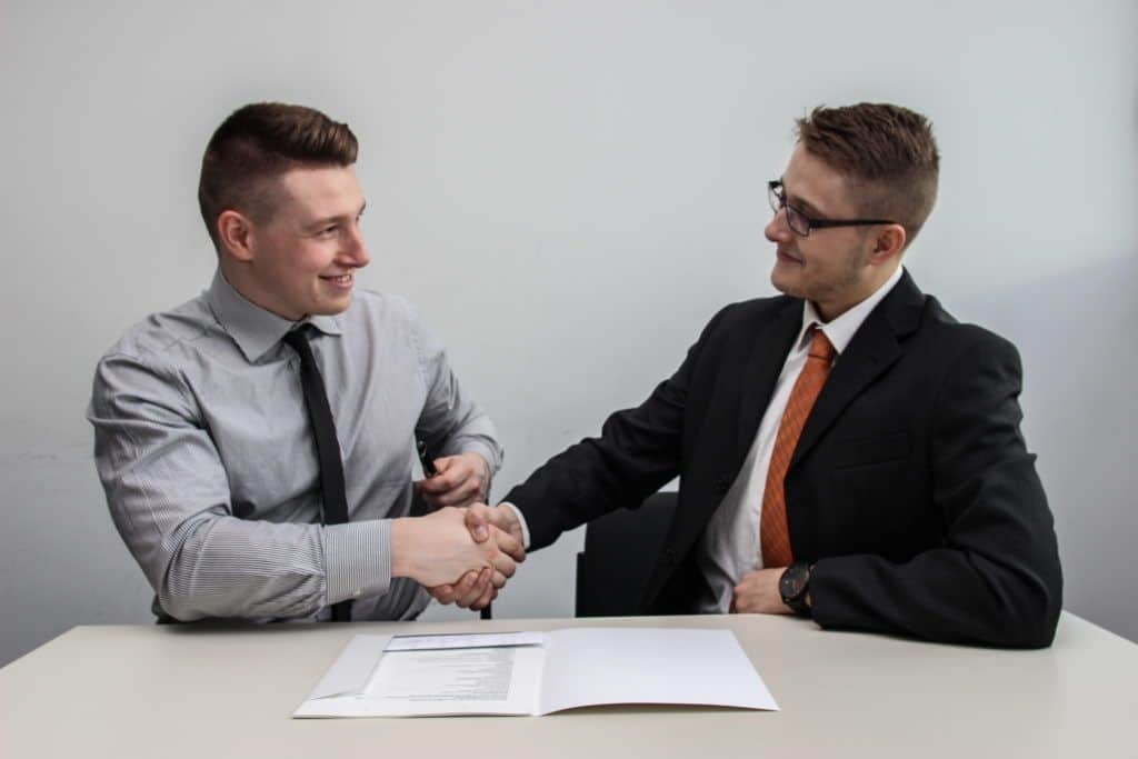 Zwei Männer im Buinesslook sitzen an einem Tisch und geben sich die Hand. Sie treffen eine Zielvereinbarung. Das Zielvereinbarungsgespräch ist ein zentrales Führungsinstrument. HR sollte es standardisiert vorbereiten und begleiten.