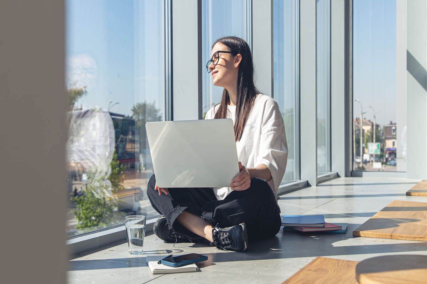 Eine Frau sitzt im Schneidersitz auf dem Boden, auf dem Schoß hält Sie ihren Laptop. Ein Symbolbild für Gleitzeit. Erfahren Sie, wie Gleitzeit im Arbeitsrecht geregelt ist und welche Vorteile und Nachteile entstehen können.